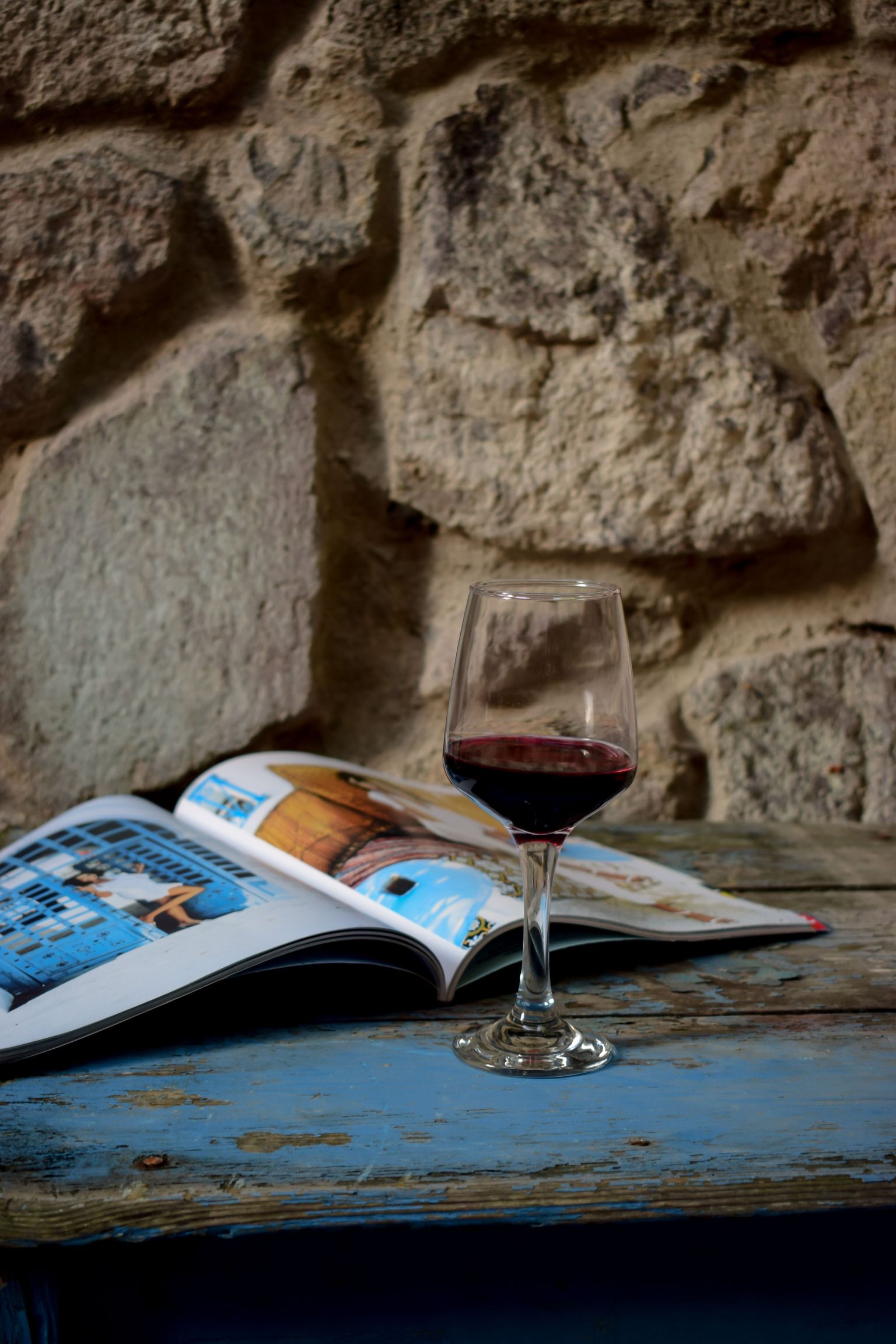 Glas mit Rotwein neben offenem Magazin auf blauer Bank vor Steinmauer.
